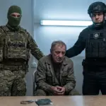 Літній чоловік у цивільному між працівником ТЦК у балаклаві та поліцейським - символ примусової мобілізації в Україні, 2026 рік