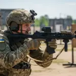 Український військовий тримає автомат, на фоні прапор України та військова техніка.