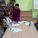 Група людей вивчає англійську мову, викладачка стоїть перед ними, у приміщенні бібліотеки.